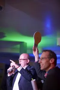 Gert-Jan Jansen met microfoon op een podium in groen en blauw licht, naast een deelnemer die een bordje omhoog houdt